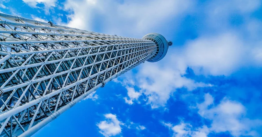 東京スカイツリーの白い鉄骨のトラス構造を真下から見上げた様子。雲の浮かぶ青空